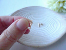 Load image into Gallery viewer, Hand holding a small gold earring against a white ceramic plate with flowers in the background